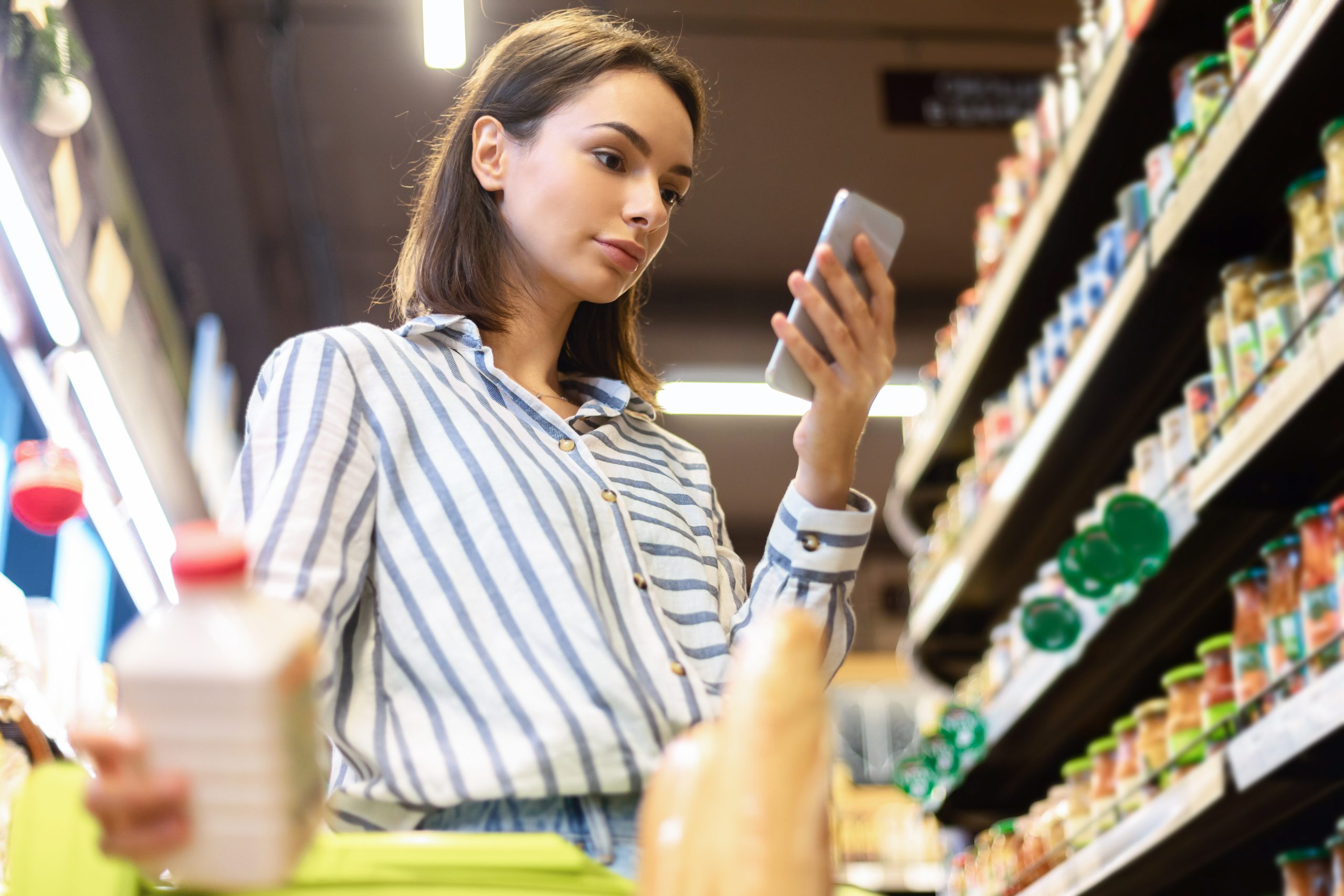 jeune femme dans un rayon de supermarché entrain de vérifier l'apport en matière grasse d'un produit sur son smartphone